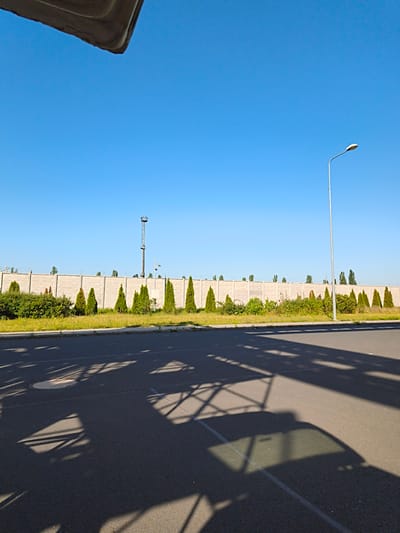 The image shows an outdoor scene with a clear blue sky. In the background, there is a long, textured concrete wall. Along the base of the wall, there are various types of greenery, including bushes and small, cone-shaped trees. A tall, narrow tower structure with a platform at the top is visible in the middle distance. To the right, a streetlamp with a curved pole stands beside the wall. In the foreground, there is an asphalt surface, likely a road or parking lot, with distinct shadows cast across it. The shadows suggest the presence of overhead structures, possibly a canopy or a large object blocking the sunlight. The scene appears to be an urban or semi-urban environment, with the concrete wall and streetlamp indicating infrastructure. The image does not depict any people.