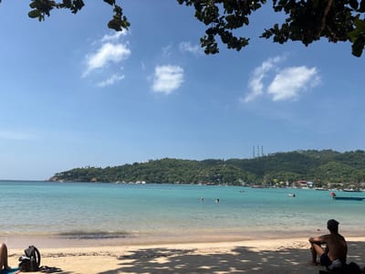 A scenic beach view showcases turquoise water lapping gently against a sandy shore. Lush green hills rise in the background, dotted with structures and topped with communications towers. Several individuals are swimming and a boat floats in the calm sea. A man sits contemplatively in the foreground shade, while another person rests further left with their belongings nearby. Tree branches frame the top of the image under a clear, blue sky with scattered clouds.