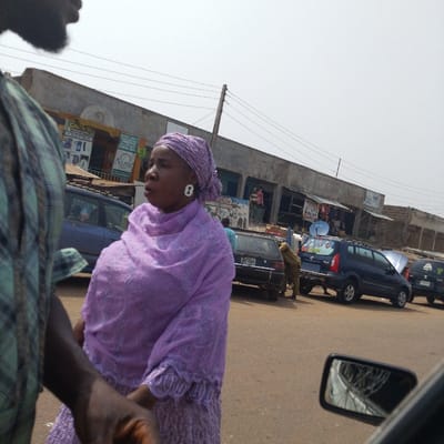 The image depicts a bustling street scene in Olomoda, Nigeria, under a bright, hazy sky. In the foreground, a partial view of a person wearing a plaid shirt is visible on the left. Centered in the mid-ground is a dark-skinned woman, facing right with her mouth slightly ajar. She wears a light purple headwrap, a matching embroidered long-sleeved shawl, and a long purple skirt, complemented by white, elongated, oval-shaped earrings.
Behind the woman, a dark blue sedan and a black sedan are visible on the reddish-brown unpaved or dirt street. Further down the street, a dark blue minivan, possibly a Mazda Premacy, is parked with its hood open, and a satellite dish is mounted on its roof; a person in a dark brown/khaki outfit attends to its rear. Another vehicle with an open hood is partially visible on the far right. A row of single to two-story commercial buildings forms the background, featuring various shop signs including "OLAS" and "Nume," with merchandise displayed behind glass. Overhead power lines and a utility pole traverse the upper frame. A vehicle's side-view mirror occupies the bottom right corner, reflecting indistinct features.