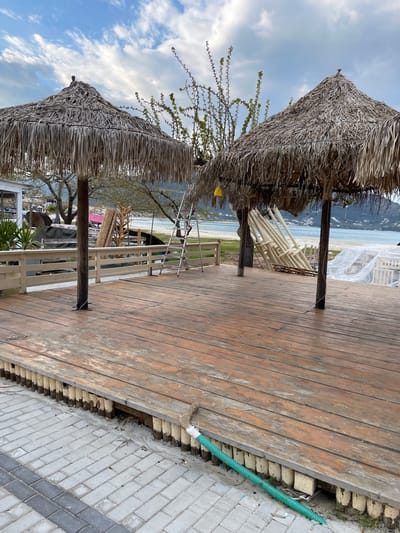 The image shows an outdoor setting in Skala Potamias, Greece. Two thatched-roof structures dominate the left side of the frame. A wooden deck occupies the central and right portions, displaying weathered planks. A green, flexible pipe runs along the edge of the deck where it meets a paved area made of light-colored bricks. In the background, a beach scene is visible, including the ocean, sand, and distant mountains. Several white objects resembling stacked chairs are behind the thatched structures. A metal ladder is standing on the deck. The sky overhead is a mix of blue patches and white clouds.