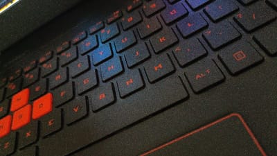 The image shows a close-up of a black laptop keyboard. The keys are black with red lettering, and the keyboard has a textured surface. The keys are arranged in a standard QWERTY layout. The image is taken from a slightly elevated angle, and the keys in the bottom left corner are in focus, with the remaining keys gradually becoming blurry towards the top right corner. The image was taken in Magelang, Indonesia, at coordinates -7.479 latitude and 110.222 longitude. The laptop keyboard is in use, with several keys depressed. The keyboard is illuminated by a light source from the top left. There is a touchpad in the bottom right corner.
