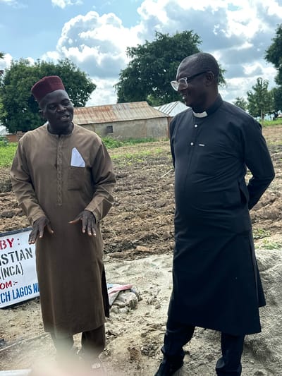 In Afogo, Nigeria, two men are standing in an outdoor, muddy field with some buildings and trees in the background. The man on the left is wearing a brown traditional African tunic with a maroon cap. He is looking to his right and speaking with the man next to him. His hands are gesturing as he speaks. To his right, a man wearing a black clerical outfit with a white collar is looking to his left and smiling. His hands are behind his back. In the foreground, a sign is partially visible with white text on a dark background. The sky above is cloudy with patches of blue.