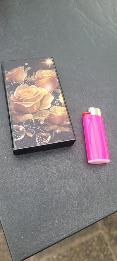 On a dark, textured outdoor surface, likely a table, two objects are visible: a cigarette case and a pink lighter. The cigarette case is rectangular and decorated with an intricate pattern of golden roses and glittering hearts. To its right, a bright pink lighter with a silver and red top is placed. The background is out of focus, suggesting an outdoor setting.