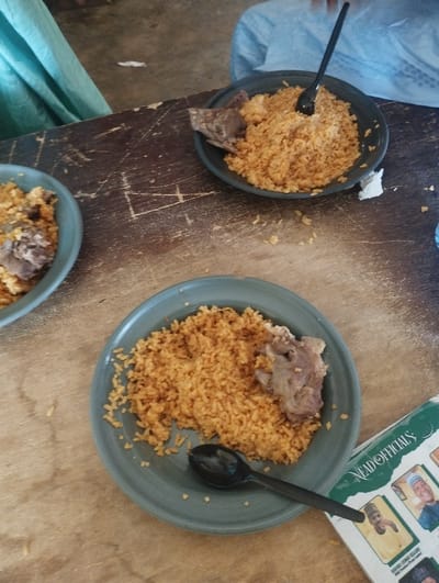 The image shows a wooden table with two plates of rice and meat. The plates are both grey and have a small amount of rice left in them. There is a black plastic spoon in one of the plates, and a black plastic spoon on the table. The table is wooden and has scratches on it. The background shows a light blue cloth and a piece of green cloth, presumably on a chair or sofa. The photo was taken at a location in Kaltungo, Nigeria, with coordinates (latitude: 9.816, longitude: 11.314). A magazine is on the table to the right of the plates, visible only from the lower right edge.  The cover of the magazine is green and includes the words “Head Officials” with several pictures of people on it.  The pictures are bordered by white and the letters are gold.