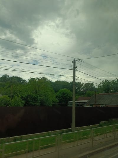 The image captures a view of a somewhat overcast sky with scattered clouds, over a residential area that appears to be in Saratovskaya, Russia. In the foreground, there's a metal fence with green grass behind it, and a dark, solid fence extending across the middle ground. Beyond the dark fence, a cluster of lush green trees forms a dense backdrop. To the right of the trees, the roofs of several houses are visible, characterized by their tiled or corrugated surfaces and chimneys. A concrete utility pole stands prominently in the mid-ground, supporting multiple overhead electrical wires that stretch across the frame. The overall impression is one of a suburban or semi-rural setting under a typical day's weather conditions.