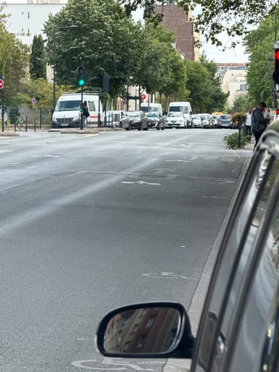 This is a photograph taken from inside a vehicle, looking out at a street scene in Colombes, France. The perspective is from the passenger side of the car, with the side mirror visible in the right portion of the frame, reflecting a building.
The street is lined with parked cars, mostly sedans and vans. The traffic signal is green, indicating that traffic can proceed. A person is visible in the background, walking on the sidewalk. Trees and buildings are present on both sides of the street. The weather appears to be overcast, with the sky being a muted grey. The overall impression is of an ordinary day in a suburban or urban environment. No discernible text is visible in the image, other than what might be on the license plates of the cars, which are not legible. The time of day is not clearly indicated, but the lighting suggests it is daytime.