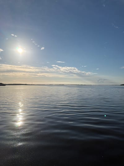 This image depicts a serene waterscape with a bright sun shining in a clear blue sky dotted with wispy clouds. The sun's reflection shimmers on the rippling surface of the water, creating a dazzling effect. In the distance, a faint landmass is visible, marked by a solitary windmill, hinting at the location of Ribnitz-Damgarten, Germany. The overall scene evokes a sense of tranquility and the peacefulness of a calm day. The time of day appears to be morning or late afternoon, given the low angle of the sun and the warm, soft light. No people or text are visible in the image.