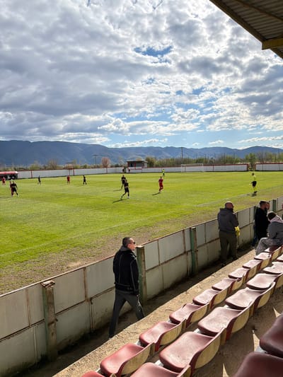 A medium shot shows a soccer match being played on a green grass field, with mountains in the background and a cloudy sky above. The field is lined with a white fence and some trees. The view is from stadium seating, where several people are watching the game, one of whom is wearing a black jacket and gray pants. The plastic seats are red. The location is most likely in Turnovo, North Macedonia.