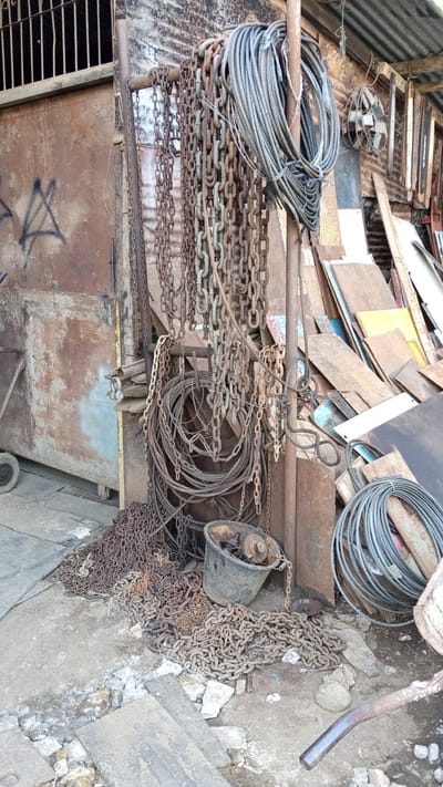 The image depicts a cluttered outdoor storage area, likely in a workshop or junkyard, located in Cotbadatunong, Indonesia. The scene is dominated by various types of heavy-duty chains, some coiled and some spread out on the ground, along with several large spools of cable. A large, rusty metal structure, possibly a building or a large piece of machinery, forms the backdrop. In the foreground, the ground is uneven, with debris, rocks, and discarded metal sheets scattered around. A dark, cylindrical container, possibly a bucket, with visible gears and a chain attached, sits near the pile of chains. The overall impression is one of disarray and industrial neglect. There are no people visible in the image. The environment suggests an area used for storing or processing industrial materials.