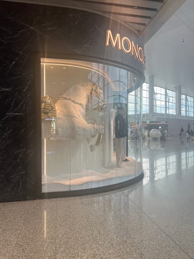 The image shows a Moncler store display window in a bright, modern airport terminal. The display features a large, white, stylized horse sculpture adorned with gold accents, possibly representing equestrian equipment. A mannequin dressed in casual attire, including a dark jacket, light-colored pants, and a cap, stands beside the horse. The store's name, "MONCLER," is prominently displayed in illuminated letters above the window.
The airport environment is characterized by polished floors that reflect the bright overhead lighting and large windows that allow natural light to stream in. In the background, other shops and restaurants are visible, including a Starbucks. People can be seen walking through the terminal, some with luggage, indicating typical airport activity. The overall scene suggests a luxurious retail space within a bustling transportation hub. The location is identified as Guangzhou City, China.