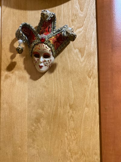 A close-up, eye-level shot captures a decorative jester mask hanging on a light-colored wooden surface. The mask, featuring a red and gold harlequin design, is adorned with small bells. The background consists of a wooden surface with visible grain and a subtle sheen, with a darker, reddish-brown vertical surface to the right, possibly indicating a doorway or cabinet edge. The lighting casts a soft shadow from the mask onto the wooden surface. The location in Yokohama, Japan is not discernible from the image.
