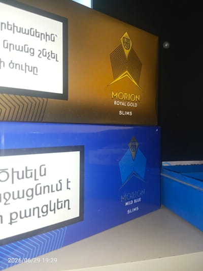 This is a close-up, slightly angled photograph of two cigarette packs, stacked one on top of the other, with a predominantly dark background and some lighter elements on the left. The lighting suggests an indoor setting, possibly a shelf or display.
The top cigarette pack is a warm golden-brown color with a stylized diamond-shaped logo featuring a geometric pattern. Text in a serif font on this pack reads "MORION ROYAL GOLD SLIMS." To the left of this pack, there is a white rectangular label with text in Armenian script. The visible Armenian text appears to be "ռեխաներին" and "Նրանց շնչել".
The bottom cigarette pack is a vibrant blue. It also features a similar stylized diamond-shaped logo. Text in a serif font on this pack reads "MORION MILD BLUE SLIMS." To the left of this pack, there is another white rectangular label with Armenian script. The visible Armenian text on this label appears to be "ծխելն" and "զգայնում է".
In the bottom left corner of the image, a date and time stamp is visible, reading "2026/06/29 19:29".
The scene does not contain any people or specific urban or natural environmental features that would allow for the identification of a location in the city of Lenina, Russia. The image is focused solely on the two cigarette packs and their labels.