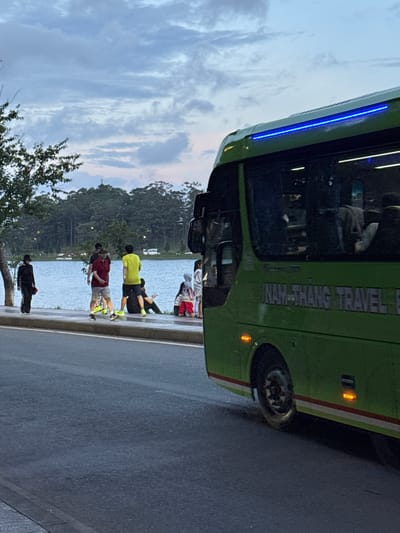 The image shows a green bus, labeled "NAM-THANG TRAVEL", parked on the side of a road next to Xuan Huong Lake in Da Lat, Vietnam. Several people are standing and sitting along the lake's edge. The sky is overcast with clouds. The scene suggests a moment of rest or observation by the lake.