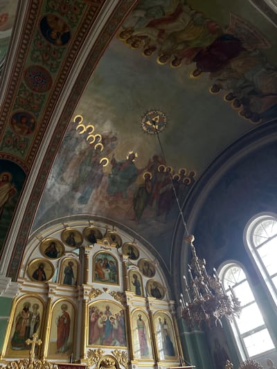 The image depicts the interior of an Orthodox church, likely located in the city of Nogornyi, Russia. The ceiling and upper walls are adorned with elaborate religious frescoes depicting biblical scenes and figures, with prominent golden halos around the heads of the figures. A large, ornate golden chandelier hangs from the ceiling, casting light downwards.
The lower part of the image reveals an iconostasis, a wall of religious icons, typically found in Orthodox churches. These icons are painted on panels and framed in gold. Various saints and biblical scenes are depicted in these icons. The overall aesthetic suggests a richly decorated and traditional place of worship. Natural light streams in from tall arched windows on the right side of the image.