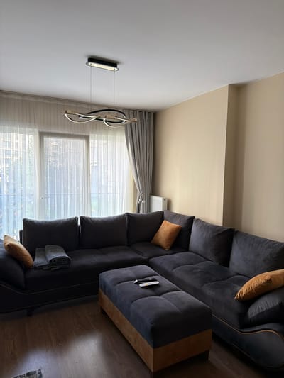 A spacious living room in Zeytinburnu, Turkey, features a large, dark grey sectional sofa with deep tufted cushions, accented by two burnt orange throw pillows and a folded grey blanket. In front of the sofa sits a dark grey ottoman with a light brown wooden base, topped with a black remote control. The room is illuminated by a modern, multi-element pendant light fixture with black and brass accents hanging from a white ceiling. Sheer white curtains cover a large window, with heavier grey drapes pulled to the sides. The walls are painted a neutral beige, and a white radiator is visible near the window. The flooring is polished dark wood.