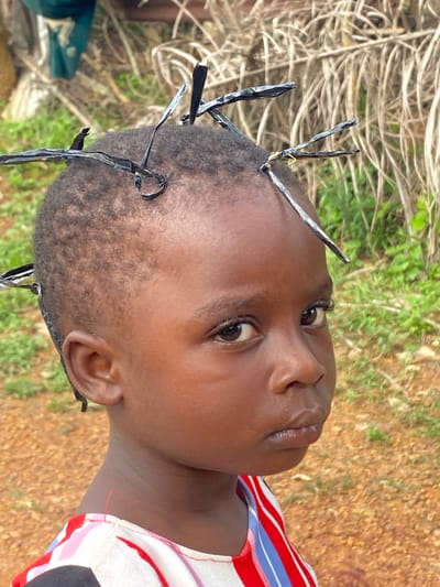 This media file features a close-up, rotated portrait of a young child with dark skin. The child's head is adorned with what appear to be thin, dark, and shiny strips of material, possibly plastic or metal, woven or secured into their hair. These strips stand out from the child's short, dark hair and point in various directions, creating a spiky effect.
The child's face is turned slightly upwards and to the right. Their eyes are open and looking off-camera, with a neutral or perhaps slightly curious expression. The lighting suggests it is daytime, likely outdoors, as indicated by the background. The background consists of dry, straw-like vegetation in the upper left and center, and reddish-brown, earthy ground with sparse green foliage in the lower right and background.
The child is wearing a colorful garment with vertical stripes of red, white, blue, and pink.
The context provided indicates the location is Ufueseni, Nigeria. No specific activity or event is discernable beyond the child being presented in this particular hairstyle. The time of day appears to be daylight, and the weather seems to be warm and dry, given the appearance of the vegetation and ground. There is no visible text in the image.
