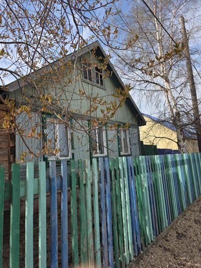 The image shows a two-story light-green wooden house in Lensk, Russia. The house has white-framed windows, some with decorative detailing. The upper story has a visible gable roof. The house is partially obscured by bare tree branches with budding leaves. Adjacent to the house, on the right side of the image, is a slatted structure made of painted wooden planks in alternating shades of green and blue. The structure appears to be a fence or retaining wall, with gravel visible behind it. The sky is visible in the background, showing blue patches interspersed with light clouds. A tree branch stretches across the top of the image.