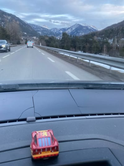 The image depicts a forward-facing view from inside a vehicle traveling on a road in the vicinity of Saint-Chaffrey, France. The immediate foreground shows the vehicle's dashboard, which exhibits extensive linear cracking across its surface. A small, red toy car, identifiable as Lightning McQueen from the "Cars" franchise, is positioned on the dashboard, facing away from the viewer.

Beyond the dashboard, a two-lane paved road (one lane in each direction) extends into the distance, marked with solid white lines along the shoulders and broken white lines in the center. Snow is present on the ground along the road's edges, and a metal guardrail lines the right side. Two other vehicles are visible ahead: a dark-colored vehicle is partially seen in the left lane, and a light-colored minivan or similar vehicle proceeds further ahead in the same lane. The environment consists of mountainous terrain with slopes covered in a mix of coniferous and deciduous trees, some with snow patches. Structures consistent with a small town or village are visible in the mid-ground among the trees. In the background, prominent snow-capped mountain peaks rise under an overcast sky.