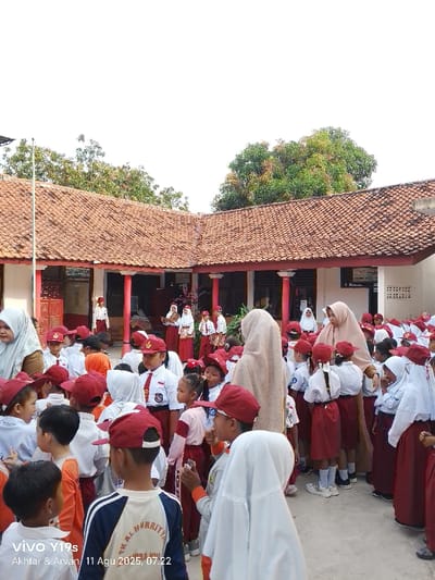 The image depicts an outdoor school scene in Jagapura Kulon, Indonesia. Approximately 50 to 70 individuals are present in a paved courtyard. The majority are children, uniformly dressed in white short-sleeved shirts, maroon skirts or shorts, and red baseball caps. Many female students wear white or light-colored hijabs. Several adult women, similarly attired in hijabs and long garments, are interspersed among the children. In the background, a single-story school building with an L-shaped, red-tiled roof and reddish-orange support pillars is visible. A tall, slender metal flagpole stands on the left side of the courtyard. Green tree foliage is discernible above and behind the building. The scene is illuminated by bright daylight.