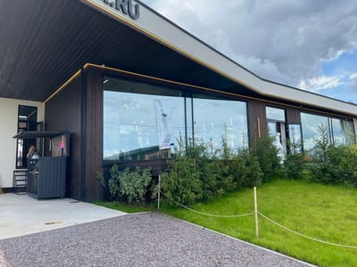 The image shows the exterior of a modern building with large glass windows and dark wooden siding, located in Yakunino, Russia. A sign with text, partially obscured, is visible on the building's facade. In front of the building, there is a paved area with gravel and concrete, and a grassy lawn with some young evergreen trees. To the left, a man is visible through a window, holding a phone. A dark gray kiosk or service booth with wheels is positioned on the paved area. The sky is cloudy with some patches of blue.