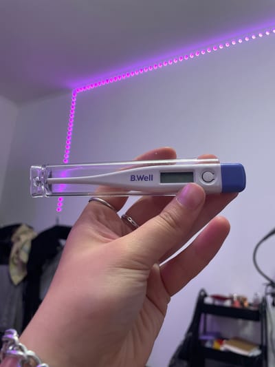 A person's hand is holding a B.Well digital thermometer in a clear plastic case. The thermometer has a white body with a blue tip and a digital display. A purple LED light strip is visible in the background, running along a wall and ceiling. The scene appears to be indoors, possibly a bedroom, with some out-of-focus objects in the background suggesting a domestic setting. The location is Vaganovo, Russia.