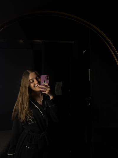 A woman in a black bathrobe is reflected in a mirror. She is holding a purple phone with stickers in front of her face and smiling. The mirror has a gold frame and is located in Ciudad López Mateos, Mexico. The environment is dark and undefined.