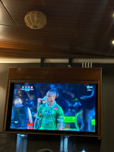 The image captures an indoor scene, likely in Saâda, Morocco, featuring a television displaying a live football (soccer) match. The television is set within a wooden frame, and the screen is rotated, showing a player in a green jersey as the primary focus.

On the TV screen, a football player, possibly of African descent, with short dreadlocks or braided hair, is prominently featured. He is wearing a bright green jersey with white trim on the collar and sleeves. Visible text on his jersey includes "Koutoubia" and "CHANGAN SINCE 1862" on the front, and "SONOP" on the sleeve. The background of the broadcast is blurred, suggesting a stadium setting with artificial lighting.

In the top left corner of the screen, a channel logo with "HD" and the Arabic word "مباشر" (Mubashir, meaning "Live") is visible. A scoreboard in the bottom left indicates the score as "0-0" and the time as "45:00". The teams are identified as "RCA" (likely Raja Club Athletic) and "RSB" (likely Renaissance Sportive de Berkane), with additional alphanumeric text "421 P2" or "421 X2" below RSB. A small, stylized 'K' or similar logo is also present in the bottom right corner of the screen.

To the left of the television, a large section of a dark brown wooden panel wall is visible. Mounted on this wall is a light-colored, woven decorative object, spherical and somewhat flattened, resembling a lantern or a basket. Behind the television frame, a light-colored pipe or conduit runs vertically along the wall, secured by dark brackets, with some white horizontal elements attached to it. The lighting in the room suggests an evening or indoor setting with artificial illumination.