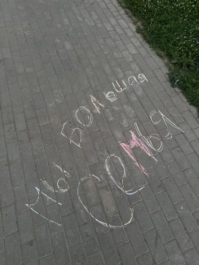 The image shows chalk drawings on a paved walkway in Mozhga, Russia. The chalk drawings, in white and pink, appear to be of letters and possibly a heart shape. Some of the text is in Russian, potentially spelling out "большая" (bol'shaya, meaning "big") and possibly "любовь" (lyubov', meaning "love") or other words. The walkway is made of grey rectangular pavers. To the right of the walkway, there is an area of green grass with small white flowers, characteristic of a park or green space. The lighting suggests it is daytime.