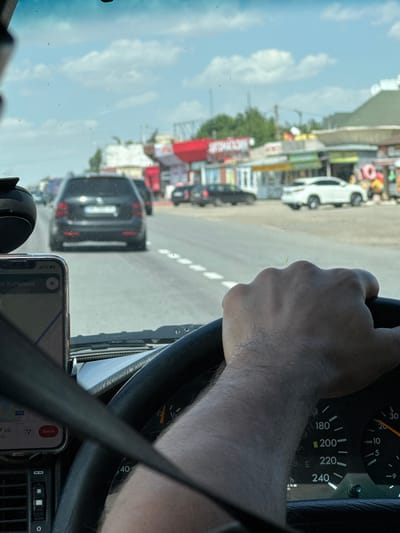 The image is taken from inside a car, looking forward through the windshield. The driver's right hand is visible, gripping the steering wheel. A smartphone mounted on the dashboard displays a navigation app showing a map and the text "ул. Котельна" (Kotelna Street), and the city of Kobleve, Ukraine. The speedometer shows a speed of approximately 80-90 km/h. Outside the car, it is a sunny day with a blue sky and some clouds. There are other vehicles on the road, including a dark SUV in front and a white car to the right. The side of the road features several small businesses and structures, characteristic of a commercial area in a town or resort.