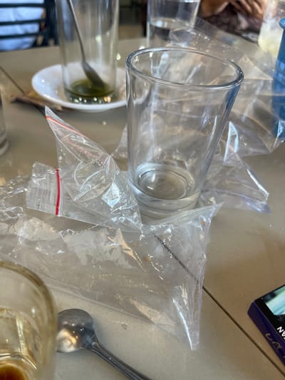 In the city of Bireuenmeunasahdayah, Indonesia, a table surface is cluttered with used drinking glasses, discarded plastic packaging, and a spoon. One glass, partially filled with a dark green liquid, sits on a white saucer. Next to it, an empty glass stands prominently. Several crumpled clear plastic bags are strewn across the table, some appearing to have been used for food or drink packaging. A metal spoon rests near the bottom left of the frame, partially obscured by a glass. In the background, a person's hands can be seen holding a patterned cloth, suggesting a dining or social setting. A blue object, possibly a bottle, is visible in the upper right corner. The overall scene depicts the aftermath of a meal or beverage consumption.