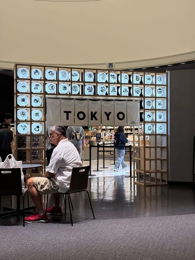 The image depicts the entrance of a store in Shinjuku, Japan, decorated with numerous illuminated Japanese lanterns featuring various traditional motifs and symbols. The lanterns are arranged in a grid structure and form an archway above the store's entrance, where a white banner with "TOKYO" in large black letters is displayed. The store interior is visible behind the banner, stocked with merchandise.

In the foreground, a man wearing a white short-sleeved shirt, khaki shorts, gray socks, and red athletic shoes is seated at a table, facing away from the camera. He has glasses and a mustache. Two dark chairs are present at the table.

To the left of the lanterns, two individuals are standing. One is a young woman with dark hair, wearing a black top and black pants. The other is a young man wearing a black shirt and dark pants.

Further inside the store, a woman in a gray hoodie and light-colored pants is walking away from the camera towards the right. Another person is partially visible behind her.

The environment appears to be an indoor public space, possibly a shopping mall or a large retail area, with polished floors reflecting the ambient light. The overall impression is one of a vibrant, culturally themed retail setting.