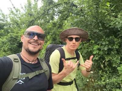 Two men are walking outdoors in a wooded area. The man on the left is bald, wearing sunglasses and a black t-shirt with a green backpack. He is smiling and looking at the camera. The man on the right is wearing a brown hat, sunglasses, and a light green t-shirt. He has a black backpack and is making a "shaka" hand gesture. The background is filled with green trees and foliage. This scene is not specific to Tazha, Bulgaria.