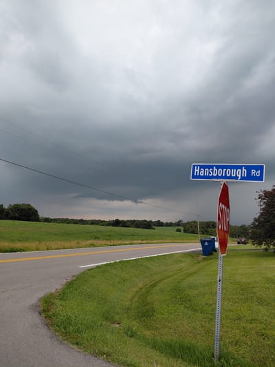 The image shows a street sign that reads "Hansborough Rd" and a stop sign. The signs are mounted on a metal pole. To the right of the signs is a blue trash bin. The road is paved with a yellow line down the center and a white line on the right edge. The road is bordered by a grassy field. In the distance, there is a line of trees. The sky is cloudy and dark, suggesting that a storm may be approaching. The scene appears to be a rural or semi-rural area. There are no people visible in the image. The time of day is unclear due to the overcast sky, but it appears to be daytime.