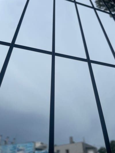 A close-up, angled view through a dark metal grate reveals a hazy, overcast sky. The grate consists of vertical bars and horizontal crossbars, creating a grid pattern. In the blurred background, elements of a city are faintly visible, including buildings with rooftops and what appears to be a large billboard or mural with blue and white imagery on the left side. The overall impression is one of a muted, possibly urban, environment seen through a barrier. The image does not provide specific details to pinpoint its location within Kołobrzeg, Poland.