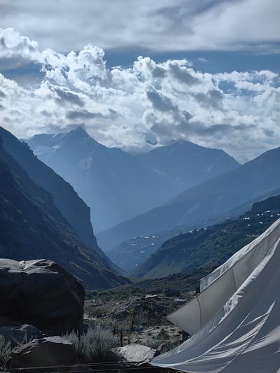 The image depicts a scenic view of the Himalayan mountain range near the village of Yamling, India. In the foreground, large rocks and dry vegetation are visible, with a section of white fabric, possibly a tent or shelter, dominating the lower right portion of the frame. The middle ground reveals a valley with a village scattered on the green slopes, characterized by small buildings and possibly tents. Further back, the majestic mountains rise, their peaks partially obscured by clouds. The sky is filled with dramatic, grey and white clouds against a bright blue backdrop. The overall impression is one of a remote, mountainous landscape with signs of human habitation nestled within nature's grandeur.