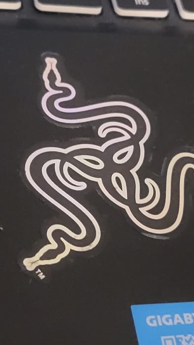 The image displays a close-up, top-down view of a metallic silver and black Razer logo sticker affixed to a matte black textured surface. The sticker features three intertwined snake heads arranged in a triangular pattern. Each snake head is distinctly outlined with pointed snouts and eye-like curves. A small "TM" trademark symbol is visible below the bottom-left snake head. The black surface, consistent with a laptop chassis, occupies the majority of the frame. Portions of three light grey keyboard keys are visible along the top edge, with the letters "ins" partially discernible on one key. A segment of a bright blue sticker with white capital letters reading "GIGABY" is visible in the bottom-right corner. The scene is situated in Magaluf, Spain. No individuals or active interactions are present.