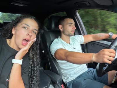 The media file shows two individuals inside a car. The subject on the left, a male, is driving and wearing a white t-shirt and grey shorts. He has a bracelet on his left wrist and a watch on his right. The subject on the right, a female, is in the passenger seat, wearing a black t-shirt and has long, dark braided hair. She is wearing a white smartwatch on her left wrist and has her hand near her mouth, which is open. Both individuals appear to be looking at the camera.

The scene is an interior car shot, with a view of greenery through the windows, suggesting they are on a road trip or driving through a scenic area. The lighting indicates it is daytime, possibly with overcast skies given the diffused light. The mood appears casual and playful, with the female subject making a surprised or excited expression.

Visible text includes "EB 5627 BH" on a tag attached to the steering wheel. The location context provided is Хаджийско, Bulgaria. The image is oriented such that the subjects are upside down.