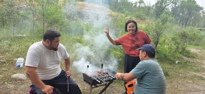In the city of Кашка-Суу, Kyrgyzstan, three individuals are gathered around a barbecue grill in a natural, grassy environment with scattered trees and a sloped, rocky hillside in the background. The scene is filled with smoke emanating from the grill, where meat skewers are cooking.
On the left, a man with a dark beard, wearing a white t-shirt and dark pants, sits facing the grill and appears to be attending to the cooking. To his right, a woman in a red t-shirt with a floral print stands and smiles at the camera, holding up two fingers in a peace sign. In front of the grill, a man wearing a blue baseball cap and a teal t-shirt is leaning over the grill, holding an orange canister, possibly for the barbecue.
An empty plastic water bottle is visible on the ground to the left of the man in the white t-shirt. The overall setting suggests an outdoor picnic or gathering.