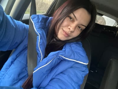 A female subject is depicted inside a vehicle, located in the city of Padarevo, Bulgaria. She is seated, with a dark seatbelt secured across her chest and left shoulder. The subject has long, dark hair and visible freckles on her face, exhibiting a slight smile while looking directly at the camera. She is wearing a bright blue, quilted puffer jacket with white trim along the collar/zipper area, over a dark garment. Her left arm is partially visible, extended towards the camera, indicating she is holding the imaging device. The vehicle's dark interior is visible, including portions of a seat and headrest. Through the vehicle's windows, a blurred outdoor background is visible, suggesting natural light and an unspecified outdoor setting.