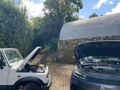 In an outdoor paved area in Vila Nova de Cerveira, Portugal, two vehicles are parked with their hoods open, revealing their engine compartments. On the left, a white, older model 4x4 utility vehicle, possibly a Suzuki Samurai, is positioned. Its hood is fully raised. On the right, a dark gray or blue modern SUV, with a prominent front grille and contemporary headlight design, is partially visible, also with its hood open. Both vehicles are situated on a surface composed of irregular stone paving. The background features dense green foliage, appearing as a tall hedge or natural growth, on the left side of the frame. To the right, behind the SUV, a building is visible, characterized by a rough-hewn stone base and a distinct white, textured, and slightly curved upper wall. The sky above is partly cloudy. No individuals or specific activities are observed.