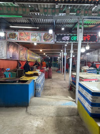 A narrow indoor space is lined with food stalls. A large LED sign displays text in a foreign language, along with food advertisements. Several people are seated along the stalls, and items for sale include bottled water and cooking oils. Gas tanks are visible near the stalls. Boxes of materials, possibly building supplies, are stacked on the right side. The setting appears to be a busy marketplace or food court.