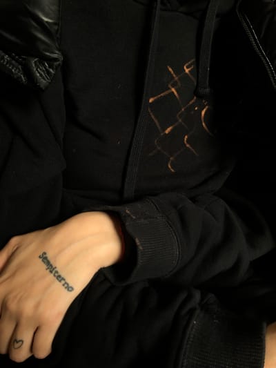 The image depicts a person wearing a black hooded sweatshirt and possibly a jacket. The sweatshirt has a pattern of what appears to be bleach stains on the front, near the drawstrings. The person's hand is visible, resting on their lap, with a tattoo on the back of the hand that says "Sempiterno" and a small heart tattoo on the thumb. The location is not determinable from the visual data provided.