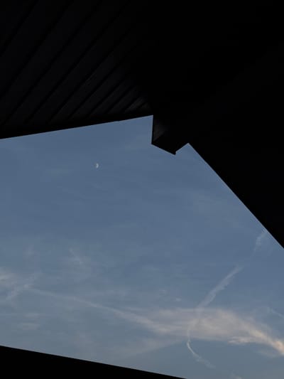This is a high-angle, close-up shot of a pale blue sky framed by dark, angular structures, likely part of a building or outdoor seating area. A sliver of the crescent moon is visible in the upper left quadrant of the sky. Wispy white clouds, some resembling contrails, are scattered across the sky. The dark structures, which appear to be made of wood or a similar material with a ribbed texture, create a stark contrast with the sky. The overall impression is one of architectural elements framing a celestial view. The image does not contain any people or specific action.