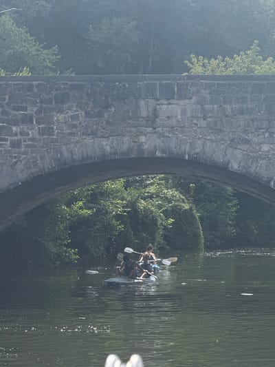 The image shows a group of people kayaking on a body of water, passing under a stone bridge. The bridge appears to be made of rough-hewn stone blocks. Lush green trees and foliage line the banks of the water. The lighting suggests it is daytime, with some atmospheric haze. The kayaks are light-colored and appear to be single-person vessels. The people are wearing casual clothing, and at least one person is wearing a life vest. The water is calm with some ripples and reflections of light on its surface. This scene is likely the River Maas in Dinant, Belgium, known for its scenic bridges and riverside activities.