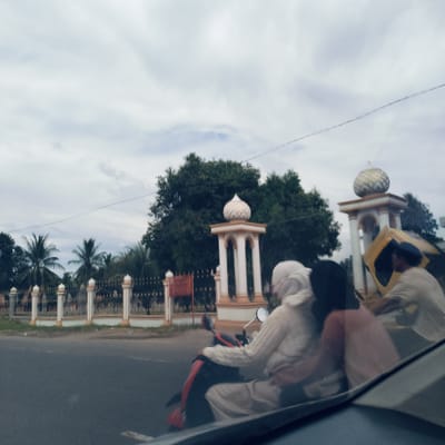 The image depicts a street scene in Gampongrayadagang, Indonesia, captured from inside a vehicle, with a partial view of the dashboard or windshield in the foreground. The sky is overcast with diffuse light, indicating a cloudy day.

The main subjects are people on motorcycles. In the midground, a dark-colored motorcycle, possibly red and black, carries at least two individuals. The front rider, seen from behind, wears light-colored clothing and a head covering, similar to a hijab. A passenger is seated directly behind them. Further to the right, another person on a motorcycle is visible, appearing to transport a large, yellowish, box-like object.

The background features distinctive local architecture, including two decorative pillars topped with white, patterned dome-like structures or cupolas. These pillars are part of a fence or gateway. Behind this structure, dense green foliage, including tropical palm trees, fills the landscape. A single power line or cable stretches horizontally across the upper part of the frame. The road is asphalt, and a low concrete wall or curb runs alongside the fence. No discernible text is visible on any signs or objects within the frame. The scene captures everyday activity and transportation in a tropical, possibly rural or semi-urban, Indonesian setting.