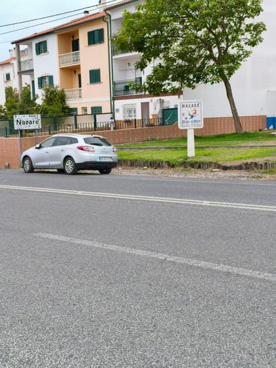 This image captures a street scene in Nazaré, Portugal. The foreground is dominated by an asphalt road with double white lines. To the left of the road, a silver station wagon is parked. Behind the car, a low brick wall and a green fence separate the road from a grassy area. In the background, there are residential buildings with light-colored facades and red-tiled roofs. A prominent feature is a white signpost with the word "NAZARÉ" at the top and "BEM-VINDO" (Welcome) below, along with "WELCOME" and "BIENVENUE" in smaller text. A large tree with green foliage stands to the right of the signpost, leaning towards the right. The sky is overcast.