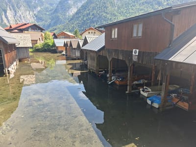The image shows a waterside view of wooden boathouses built on stilts over a body of water in Obertraun, Austria. The boathouses are made of weathered wood, with some featuring metal roofs. Several small boats are docked inside or near the boathouses, including at least two motorboats and a pedal boat. The water is clear in some areas, revealing the sandy bottom, and dark in others, reflecting the structures and surrounding mountains. The backdrop features a steep, tree-covered mountainside with some buildings visible on its lower slopes. A sign with German text is attached to one of the boathouses. The overall scene suggests a calm, possibly summer, day with typical lakeside or fjord activity.