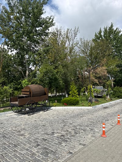 The image depicts an outdoor scene with a cobblestone pathway leading through a park-like setting. In the foreground, a vintage wooden wagon with a large barrel and seating is parked on the cobblestones. Further back, a vintage military truck is partially visible among the trees. The environment is lush with greenery, including various trees and bushes, and the sky above is cloudy. Two orange and white traffic cones are placed along the edge of the pathway. The location is identified as the city of Mirikənd, Azerbaijan. There are no people visible in the image.