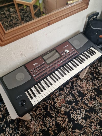 A Korg Pa700 Professional Arranger keyboard sits on a wooden stand over a patterned carpet. The keyboard is dark grey and maroon with a display screen and numerous buttons. To the left of the keyboard, a speaker grille is visible. A wooden framed mirror with a reflection of a wooden chair and a floral carpet is situated above and behind the keyboard. A black bag with grey trim leans against a textured wallpaper wall to the right of the keyboard. The scene appears to be indoors, likely in a home setting in King's Lynn and West Norfolk, United Kingdom.