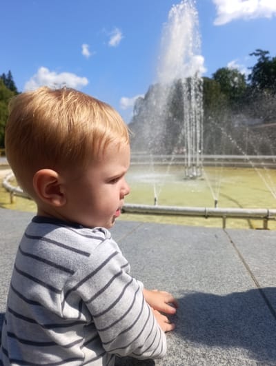 A young child with blonde hair, wearing a grey and black striped shirt, sits on a granite ledge and looks to the right. Behind the child, a fountain sprays water into the air, with trees and a blue sky visible in the background. The scene appears to be outdoors, possibly in a park or public square, given the fountain and paved area. The location is Luhačovice, Czechia.